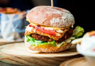 Close up image of a freshly flame grilled vegetarian halloumi cheeseburger on a wooden counter at an outdoor food market. This burger is loaded with a vegetarian burger meat substitute, fresh salad, melted halloumi cheese and spanish onion and tomato. The burger is sandwiched between glazed buns.